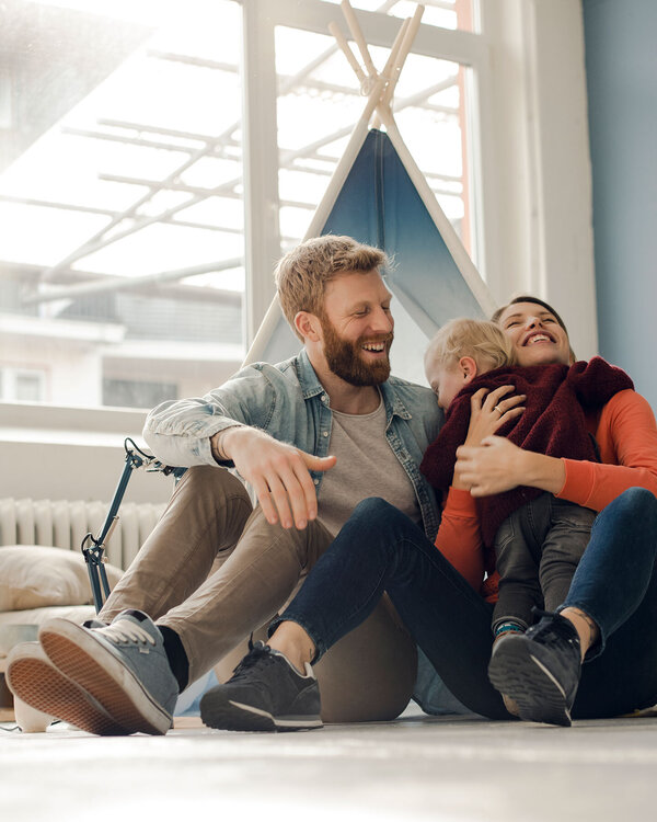 Familie mit Kleinkind sitzt im Kinderzimmer am Boden und umarmen das Kind